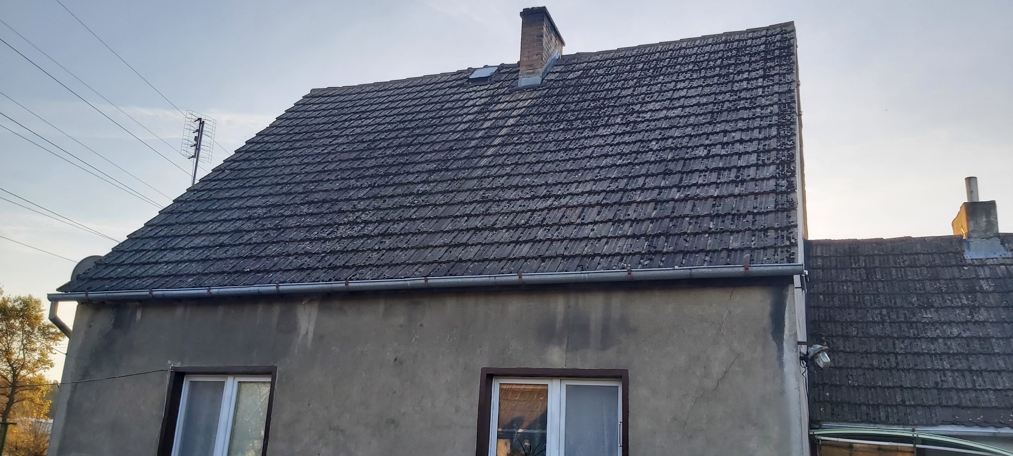 Stary dach z dachówki ceramicznej pokryty mchem, widoczny komin, rynna i antena telewizyjna na tle nieba.