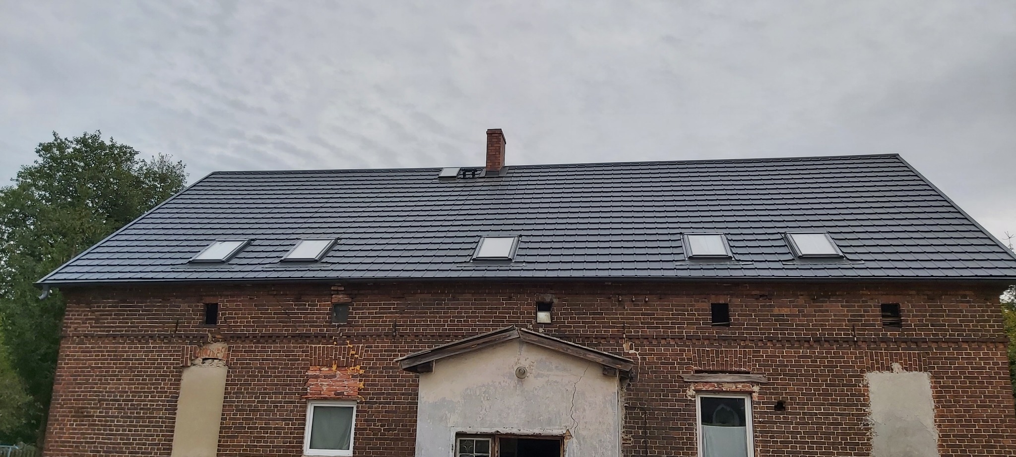 Szary dach z dachówką bitumiczną na ceglanym budynku z oknami dachowymi i kominem.