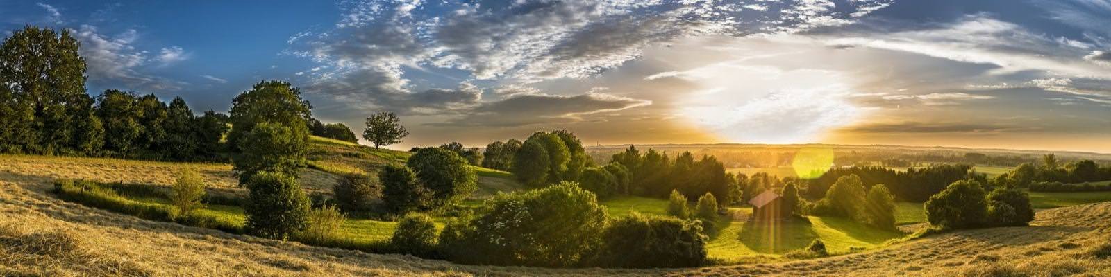 Panoramiczny widok na wiejski krajobraz o zachodzie słońca, z polami, drzewami i budynkiem w oddali, pod błękitnym niebem z chmurami.