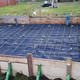 Zbrojenie fundamentu na folii, widoczna drewniana szalunek z zielonymi wspornikami, pracownik w ciemnej kurtce dogina pręty zbrojeniowe, w tle trawnik i ogrodzenie z siatki.