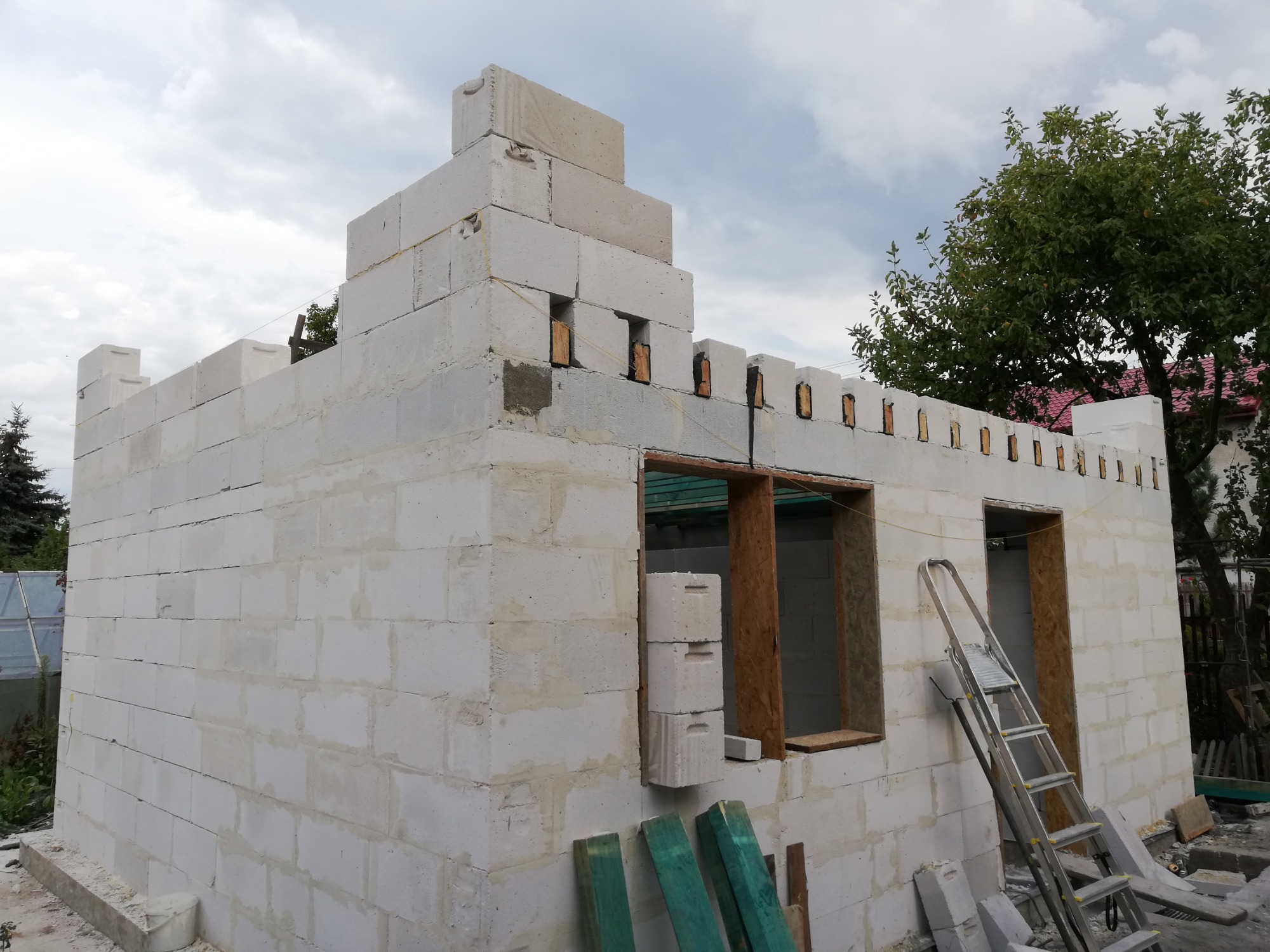 Niedokończona budowa domu z białych bloczków, widoczne otwory okienne z drewnianą ramą, drabina oparta o ścianę, drewniane deski na pierwszym planie, budynek w stylu zamku.
