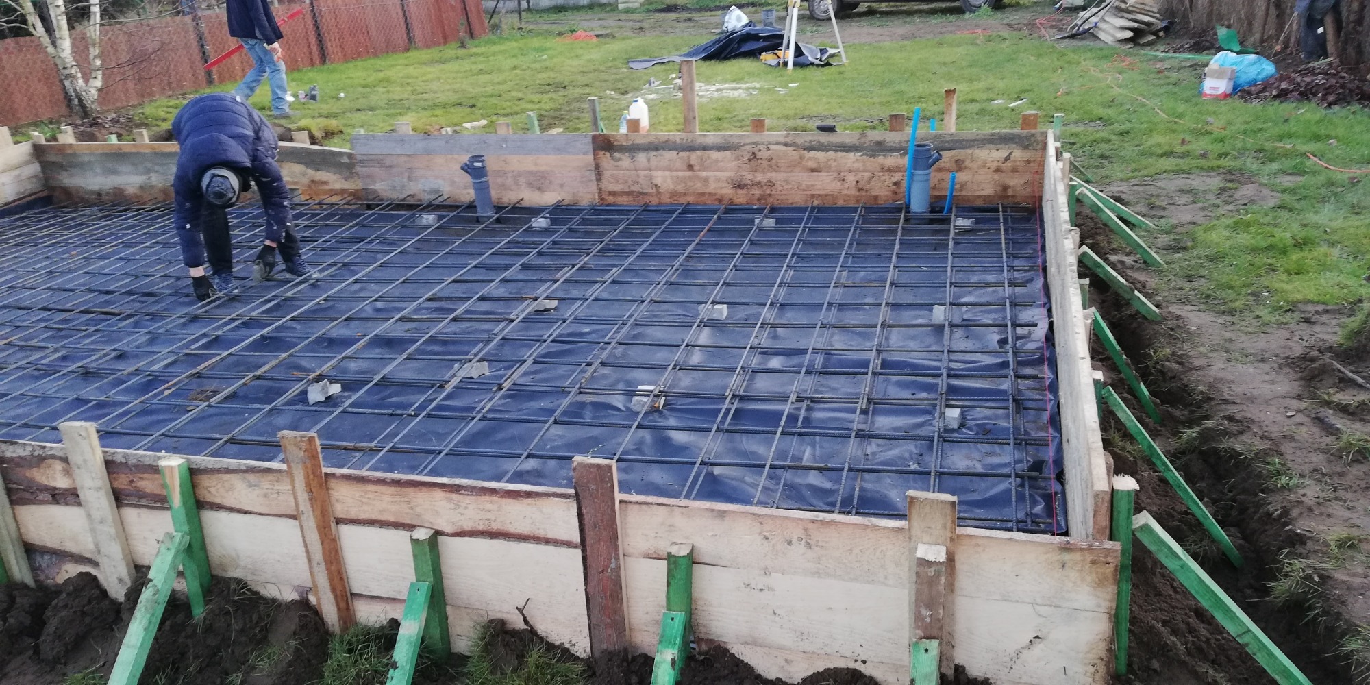 Zbrojenie fundamentu na folii, widoczna drewniana szalunek z zielonymi wspornikami, pracownik w ciemnej kurtce dogina pręty zbrojeniowe, w tle trawnik i ogrodzenie z siatki.