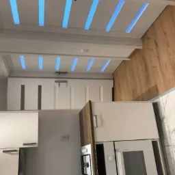 Nowoczesna kuchnia z białymi szafkami, drewnianym blatem i podświetlanym sufitem z niebieskimi paskami LED, widok z dołu.