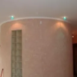 Zaokrąglona ściana w salonie wykończona dekoracyjnym tynkiem w odcieniach beżu, z wbudowanymi luksferami i oświetleniem punktowym w suficie.