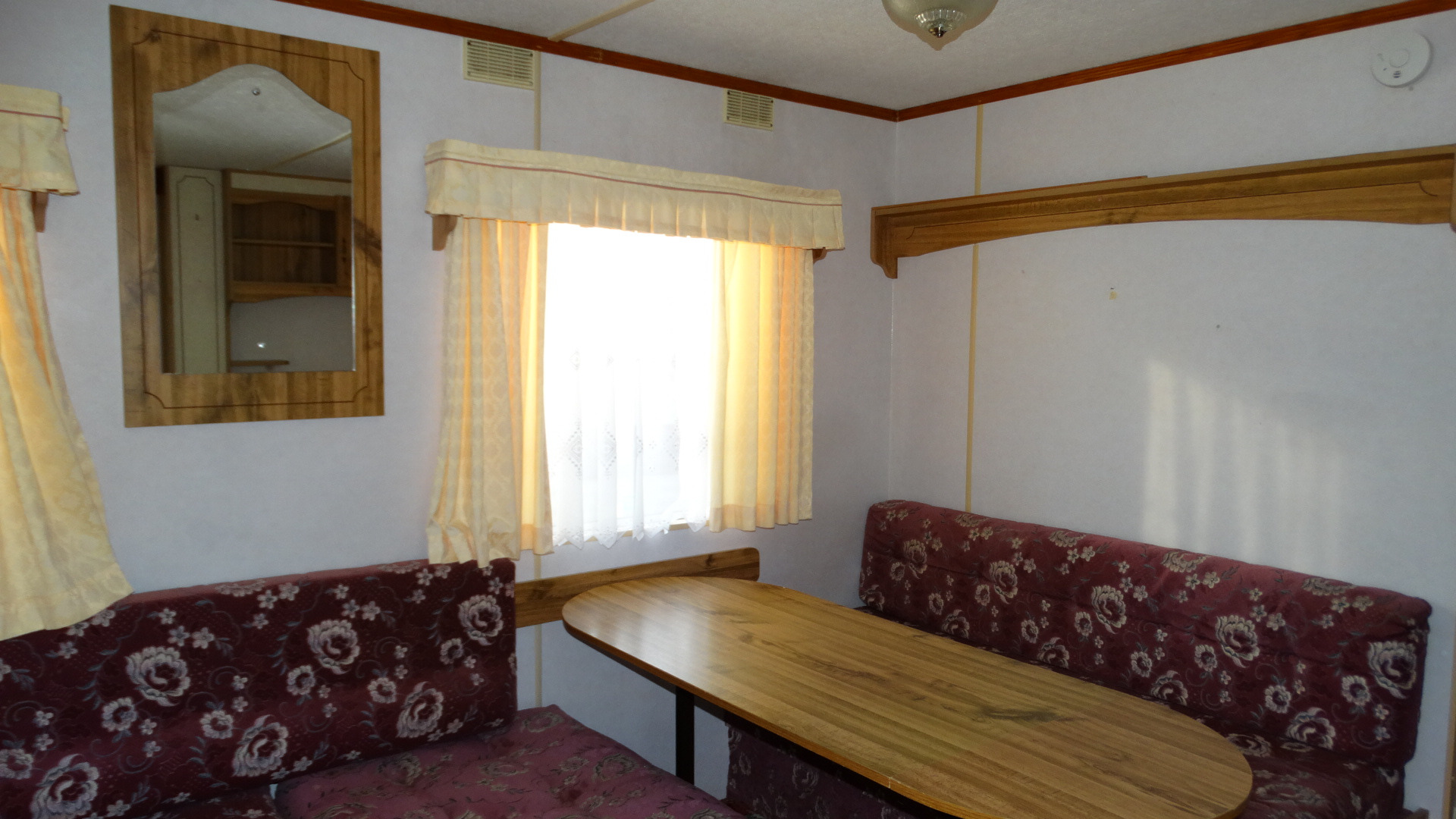 Wnętrze domku holenderskiego z drewnianym stołem, tapicerowanymi siedziskami w kwiaty, oknem z kremowymi zasłonami i lustrem na ścianie.