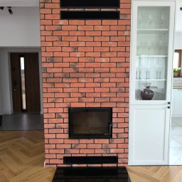 RKW Kominki Wojna Radosław - Murowany kominek Defro Intra S Mini z czarną szybą i obudową z cegły klinkierowej, wkomponowany w nowoczesne wnętrze z parkietem w jodełkę i białą zabudową.