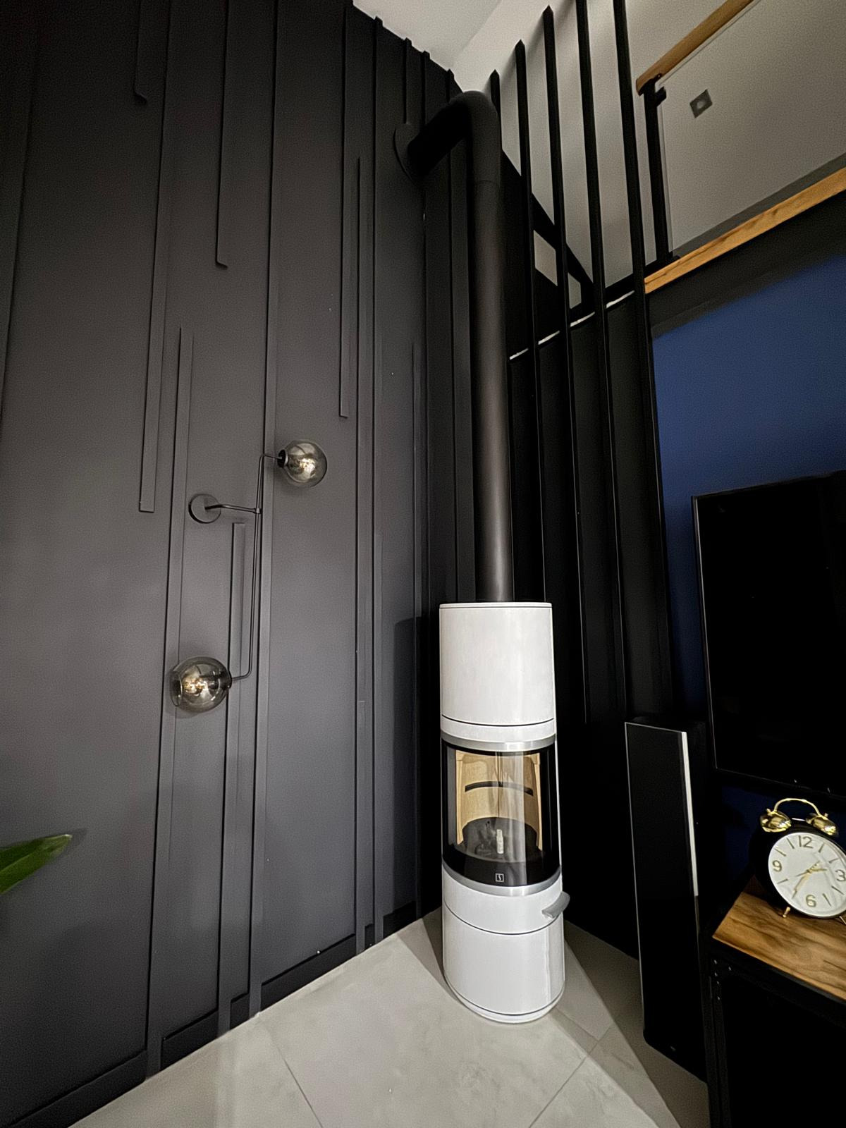 Nowoczesny piec wolnostojący w białej obudowie z czarnym przewodem kominowym, na tle ściany z dekoracyjnymi pionowymi panelami i designerską lampą kulową.