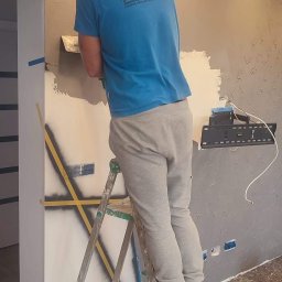 DH Daniel Haberski - Mężczyzna w niebieskiej koszulce z logo firmy Danio-Bud na drabinie, nakłada szpachlą dekoracyjny tynk na ścianę w pokoju, widoczne zabezpieczenia folią i taśmą malarską.