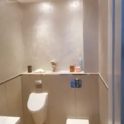 Nowoczesna, beżowo-szara toaleta z podwieszaną muszlą klozetową i pisuarową, dekoracyjna ściana z efektem chmur, półka z ozdobami i oświetleniem punktowym.