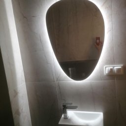 DH Daniel Haberski - Nowoczesna umywalka z szafką i podświetlanym lustrem o nieregularnym kształcie na tle marmurowej ściany z białymi żyłami, odbicie ręki z telefonem w lustrze.