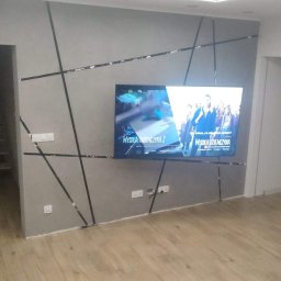 DH Daniel Haberski - Szara ściana z dekoracyjnymi czarnymi listwami tworzącymi geometryczny wzór, na której wisi telewizor. Widoczna podłoga z paneli, fragment schodów i przejście do innego pomieszczenia.