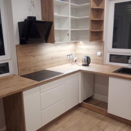 DH Daniel Haberski - Nowoczesna kuchnia z białymi szafkami i drewnianym blatem, czarna płyta indukcyjna, czajnik elektryczny na blacie, drewniana podłoga, widoczne kable wystające ze ściany.