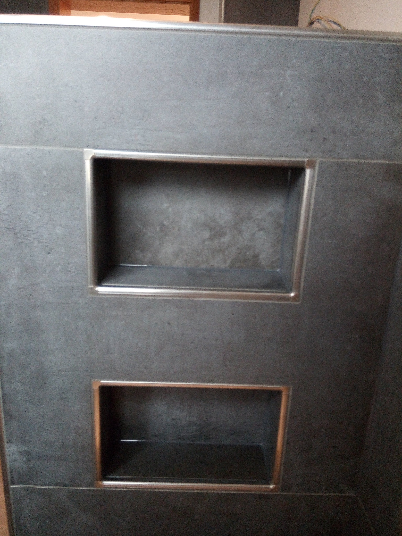 Dwa wnęki w ścianie wyłożonej szarą, matową płytką, obramowane metalowymi listwami, w górnej wnęce widoczny cień, w dolnej wnęce odbicie światła.