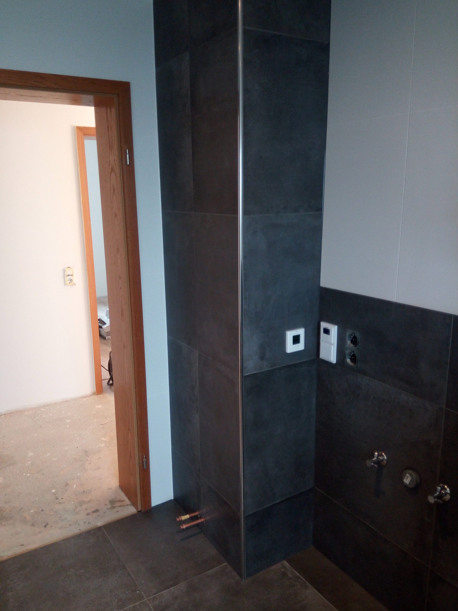 Ciemne płytki imitujące kamień, ułożone na ścianie i podłodze w łazience, z widocznym fragmentem instalacji wodnej i otwartymi drzwiami do innego pomieszczenia w trakcie prac wykończeniowych.