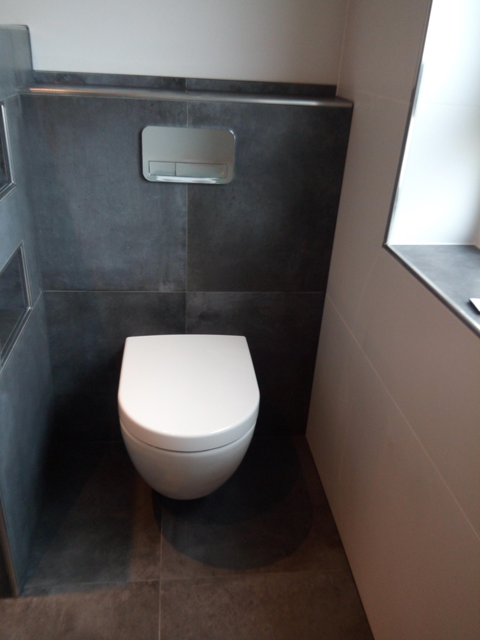 Kompaktowa toaleta podwieszana z chromowanym przyciskiem spłukującym, otoczona ciemnoszarą płytką imitującą kamień, z wnęką w ścianie i jasną okładziną po prawej stronie.