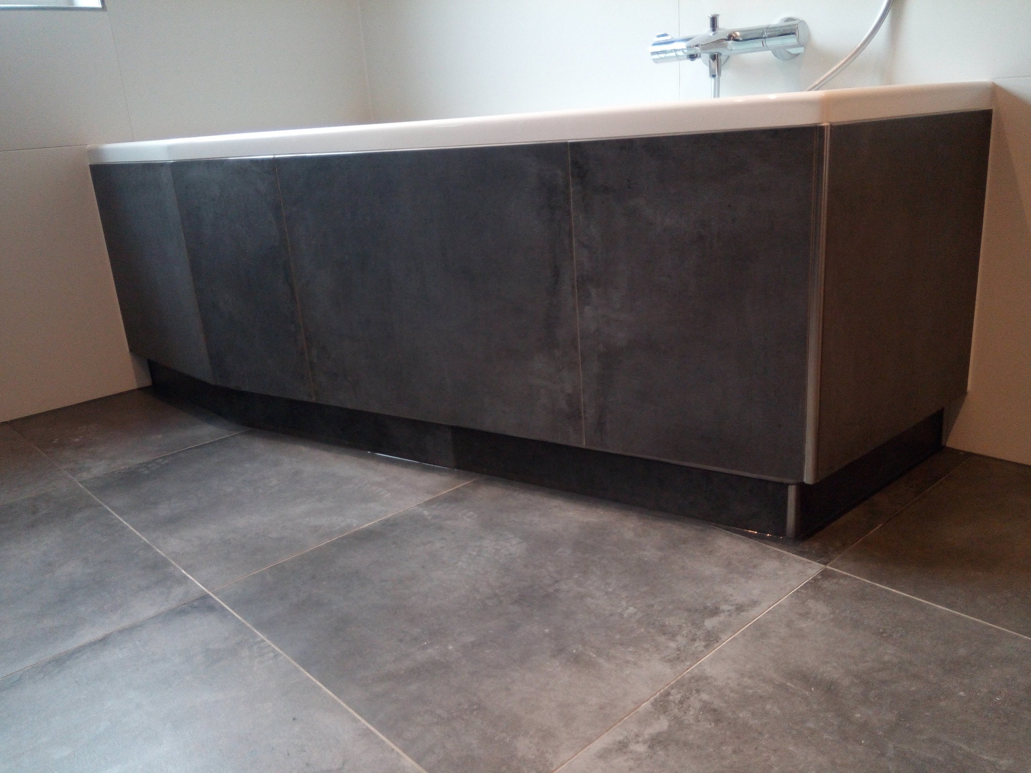 Obudowa wanny wykończona ciemnymi płytkami imitującymi kamień, dopasowana do szarej podłogi z płytek o podobnej fakturze, widoczna bateria wannowa.