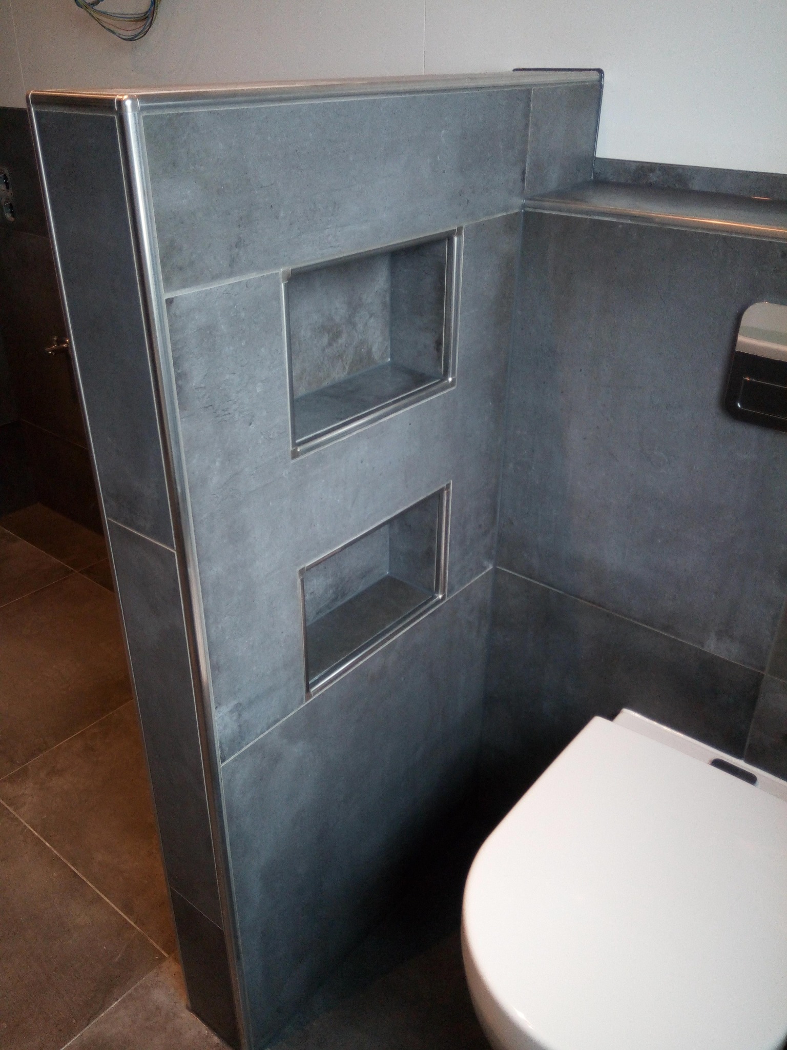 Szare płytki wielkoformatowe z wnękami wykończonymi listwami aluminiowymi w nowoczesnej łazience, widoczny fragment podwieszanej toalety.