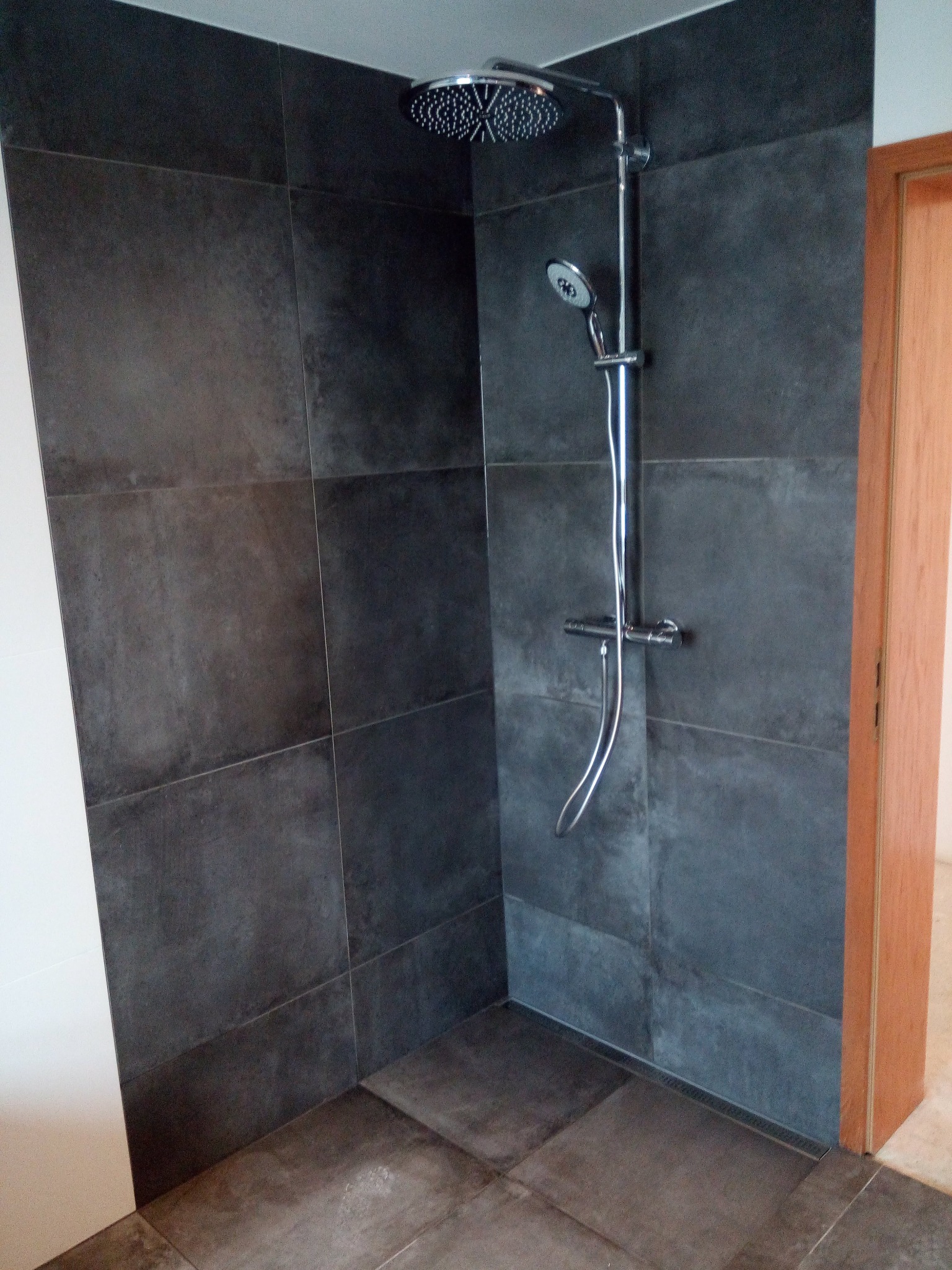 Nowoczesny prysznic z ciemnoszarą, matową glazurą i chromowaną armaturą, widoczny fragment drewnianej ościeżnicy drzwiowej.
