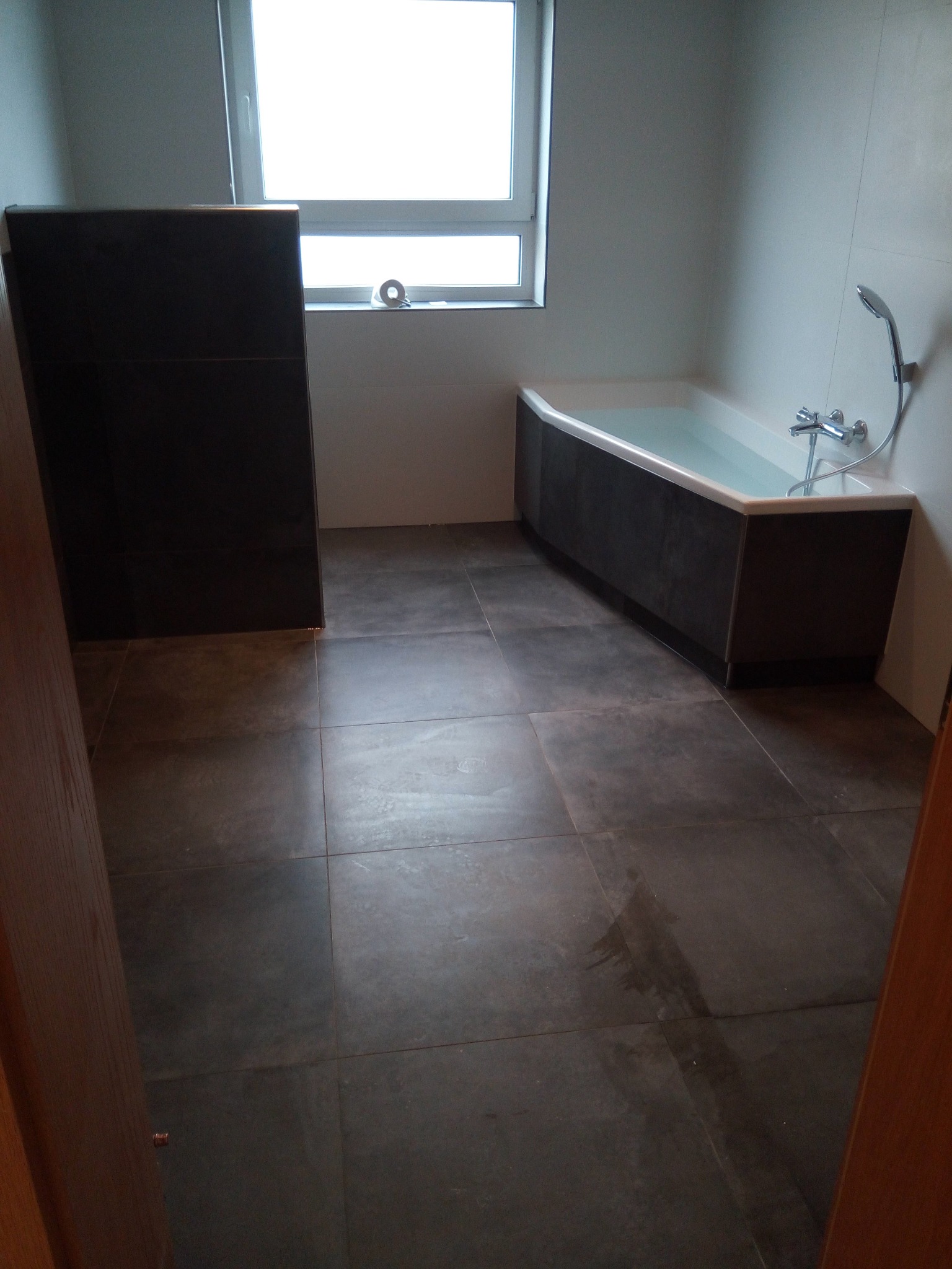 Nowoczesna łazienka z ciemnymi płytkami podłogowymi i obudową wanny, biała wanna wypełniona wodą, okno z rolką taśmy klejącej na parapecie.