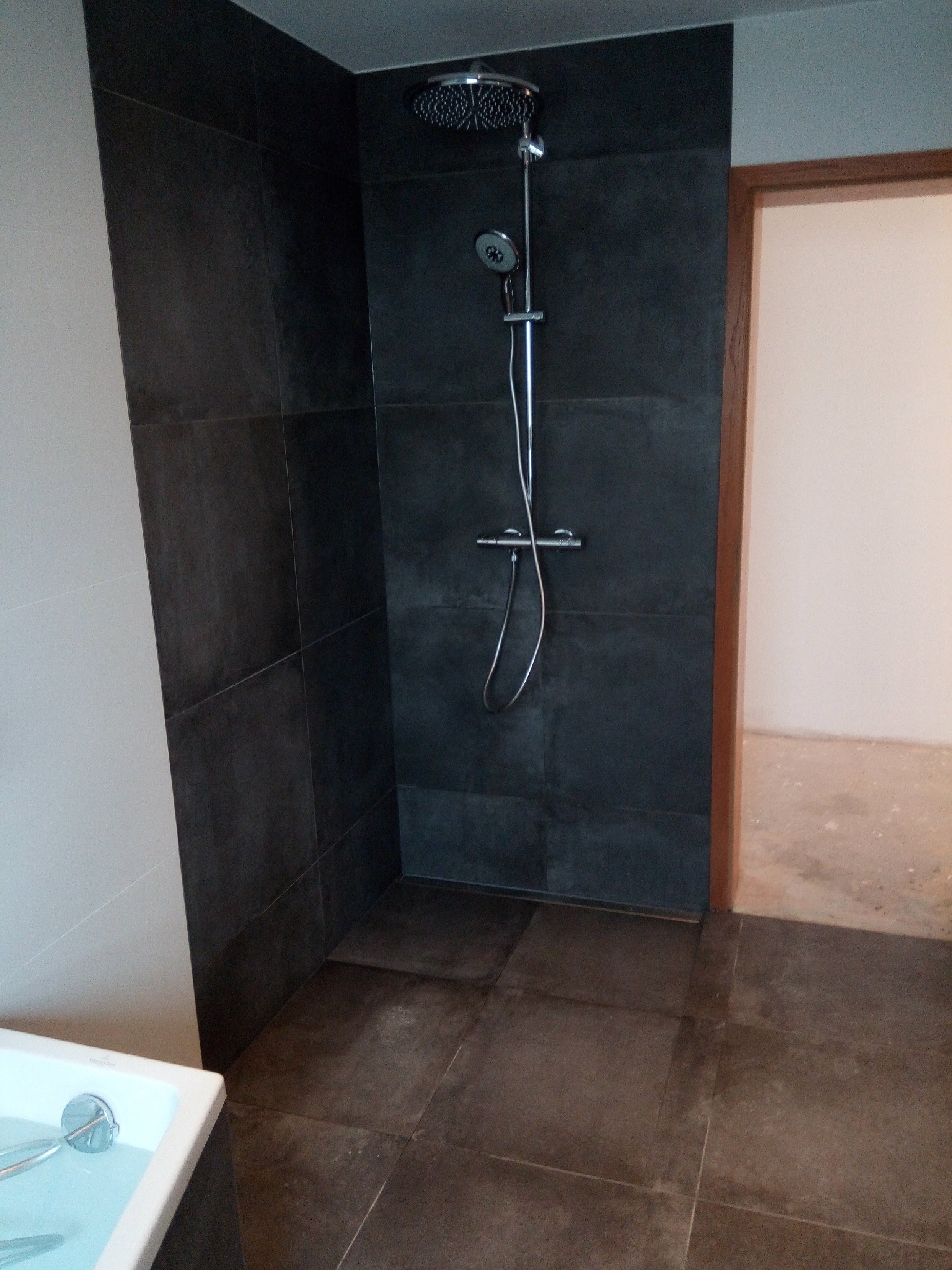 Nowoczesna kabina prysznicowa z deszczownicą i ciemnymi płytkami w trakcie montażu, widoczny fragment wanny i niedokończona podłoga w pomieszczeniu obok.