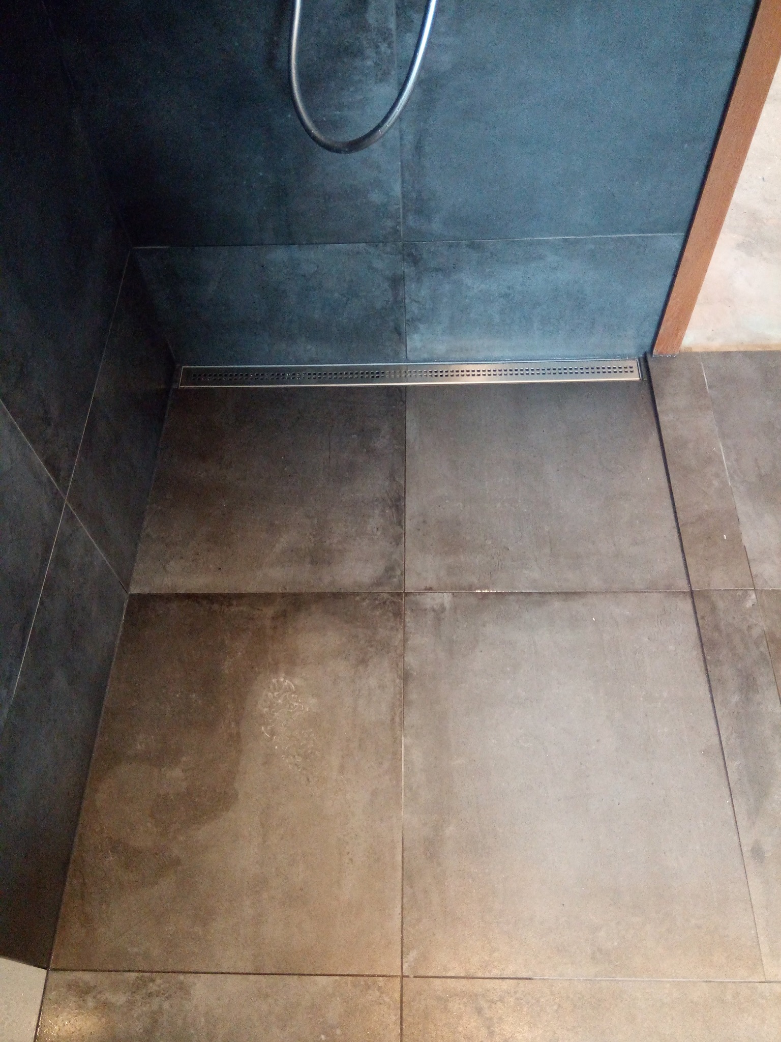 Nowoczesny odpływ liniowy w kabinie prysznicowej wyłożonej ciemnymi płytkami, widok z góry, z fragmentem słuchawki prysznicowej.