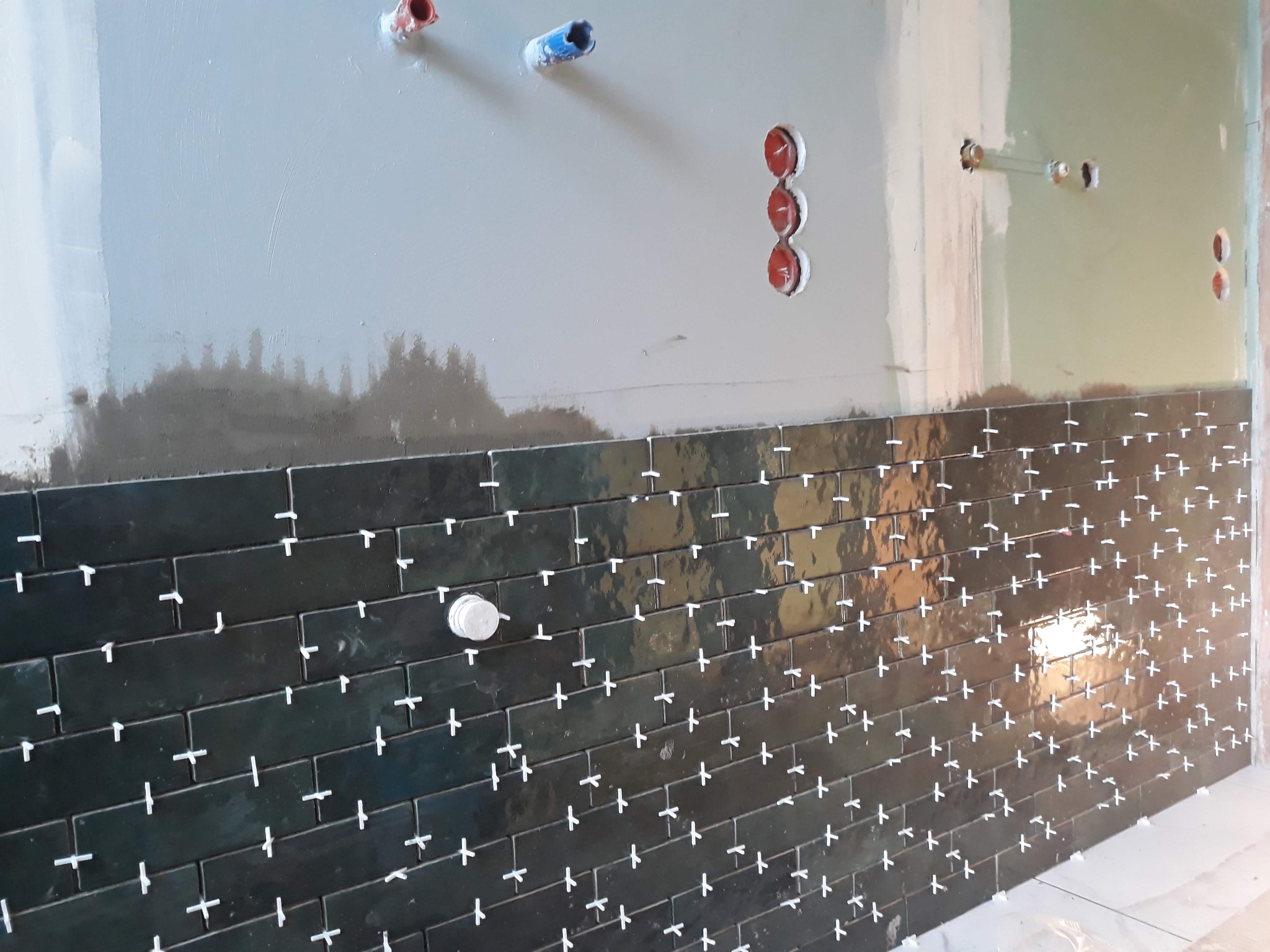 Świeżo ułożone, błyszczące, ciemnozielone płytki w kształcie cegiełek na ścianie z widocznymi krzyżykami dystansowymi, surowa ściana powyżej z instalacją wodną i elektryczną.