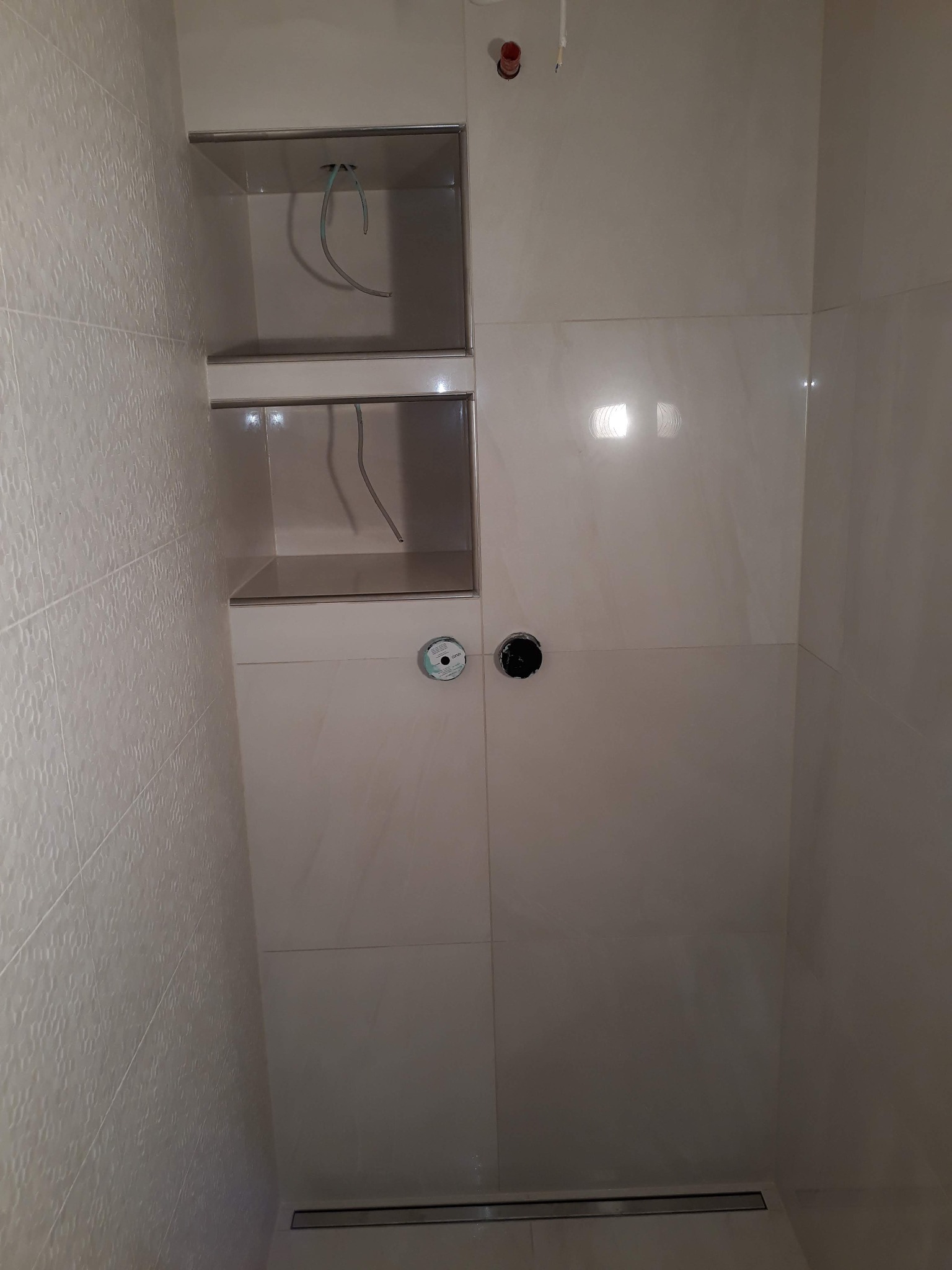 Wnętrze kabiny prysznicowej w trakcie prac wykończeniowych, widoczne dwie wnęki w ścianie z wyprowadzonymi przewodami, armatura pod prysznic i odpływ liniowy w podłodze.