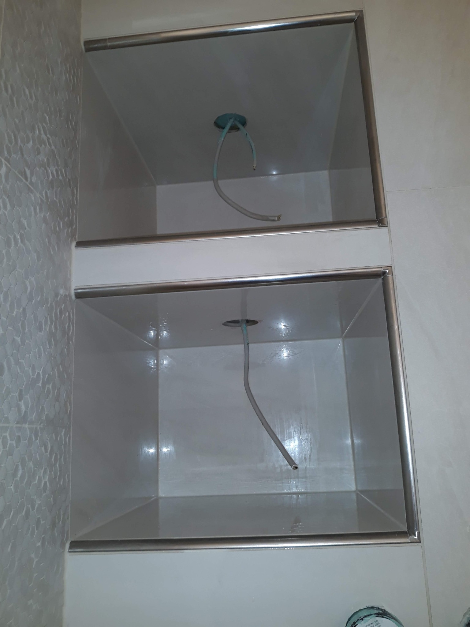 Dwie wnęki w ścianie łazienki wyłożone białymi płytkami z metalowymi obramowaniami, widoczne wystające przewody instalacji oświetleniowej.