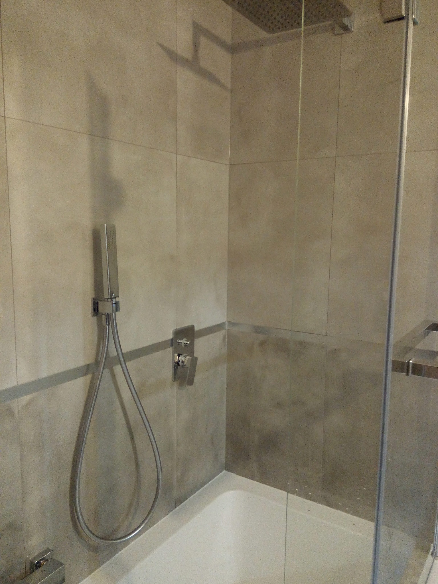 Nowoczesna kabina prysznicowa z deszczownicą i panelem sterującym, wykończona płytkami w odcieniach szarości z delikatnym połyskiem. Widoczne elementy armatury w kolorze chromu i fragment wanny.