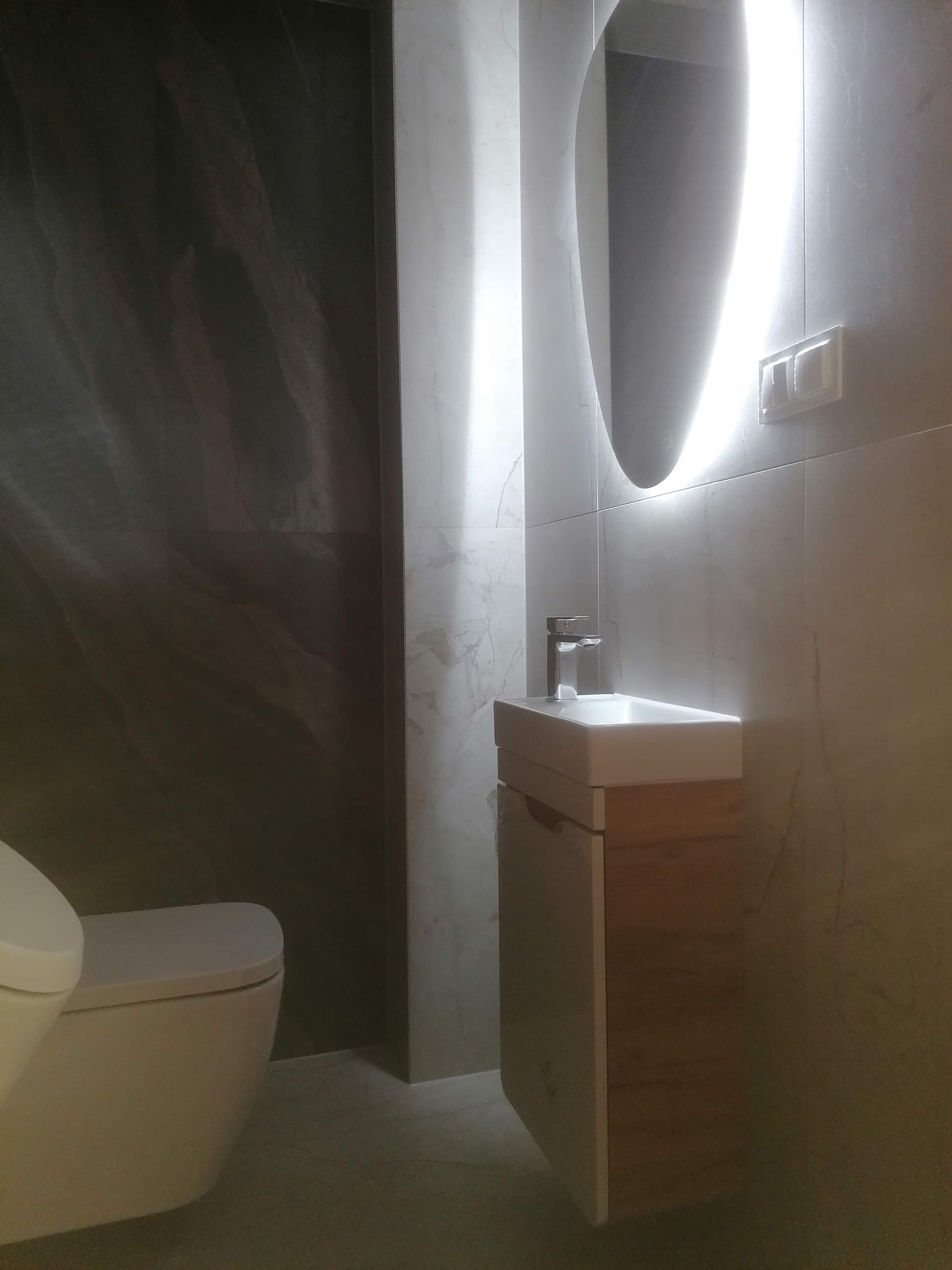 Nowoczesna łazienka z podświetlanym lustrem owalnym, umywalką nablatową na szafce z drewnianym frontem oraz toaletą w tle, wykończona płytkami imitującymi marmur i kamień.