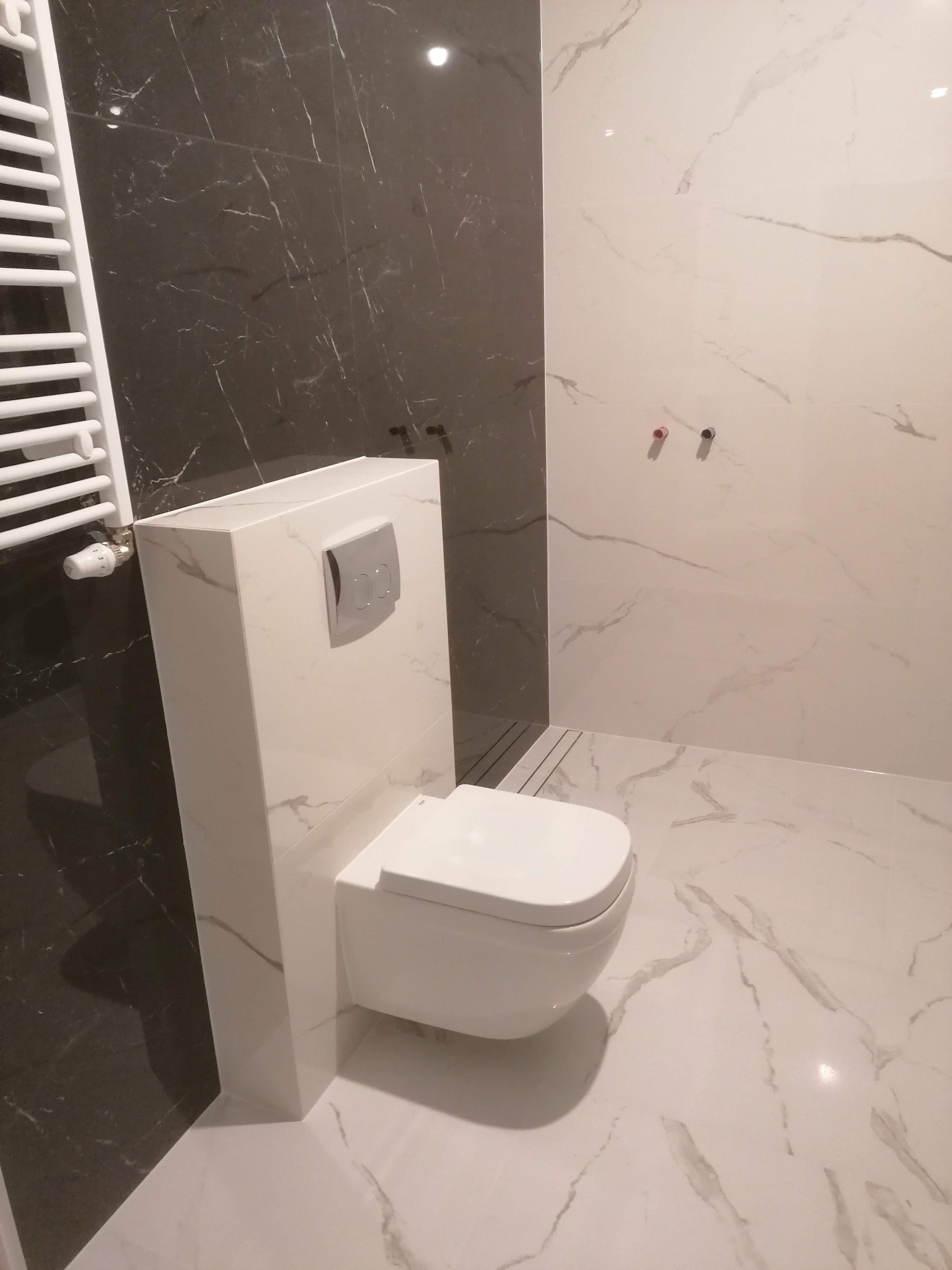 Nowoczesna łazienka z wiszącą toaletą o marmurowym wykończeniu, grzejnikiem drabinkowym i płytkami imitującymi marmur w odcieniach bieli i szarości.