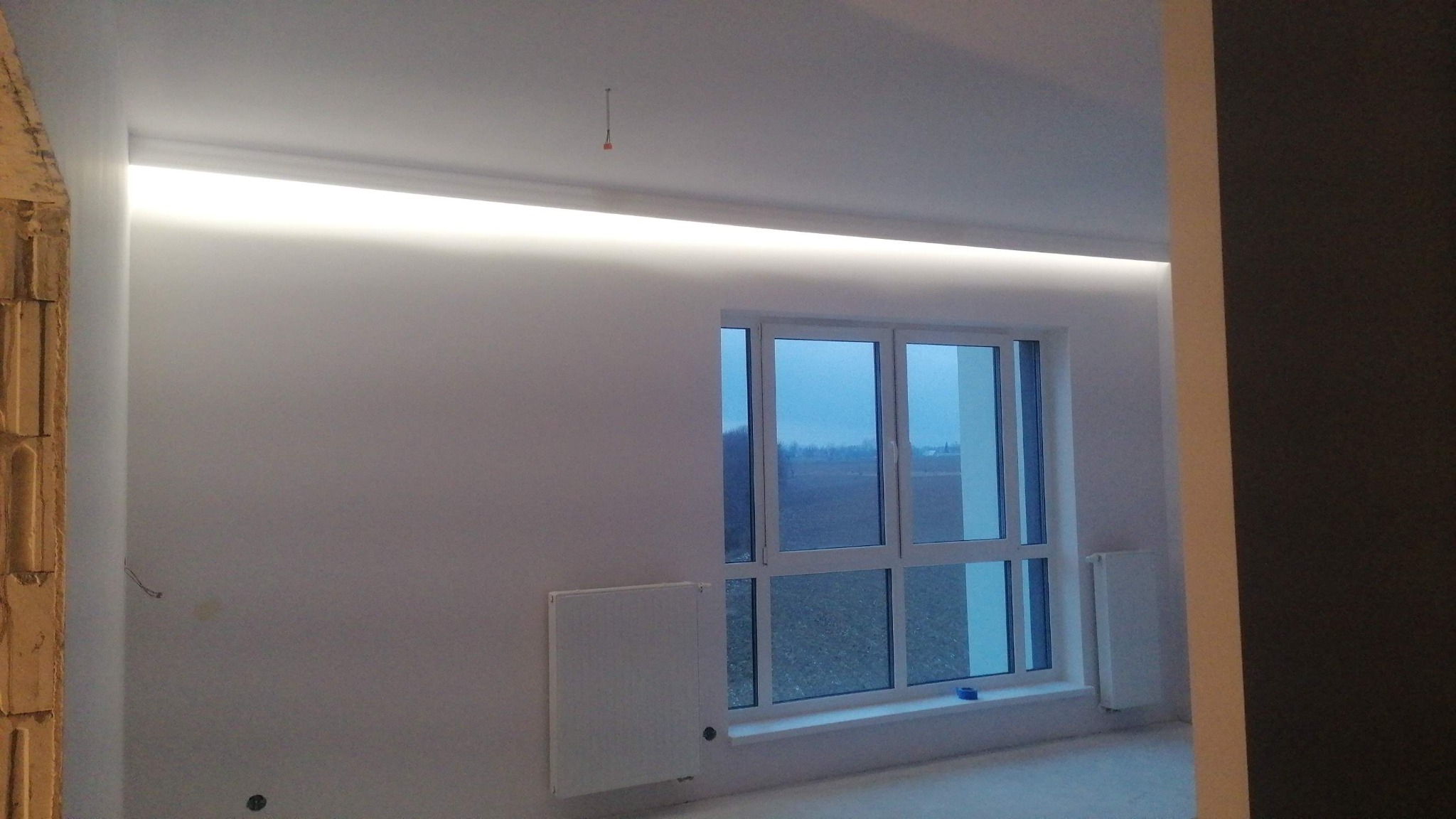 Pokój z białymi ścianami i oknem, widoczna listwa LED pod sufitem, grzejnik i fragment ściany w innym kolorze, surowy stan wykończenia.