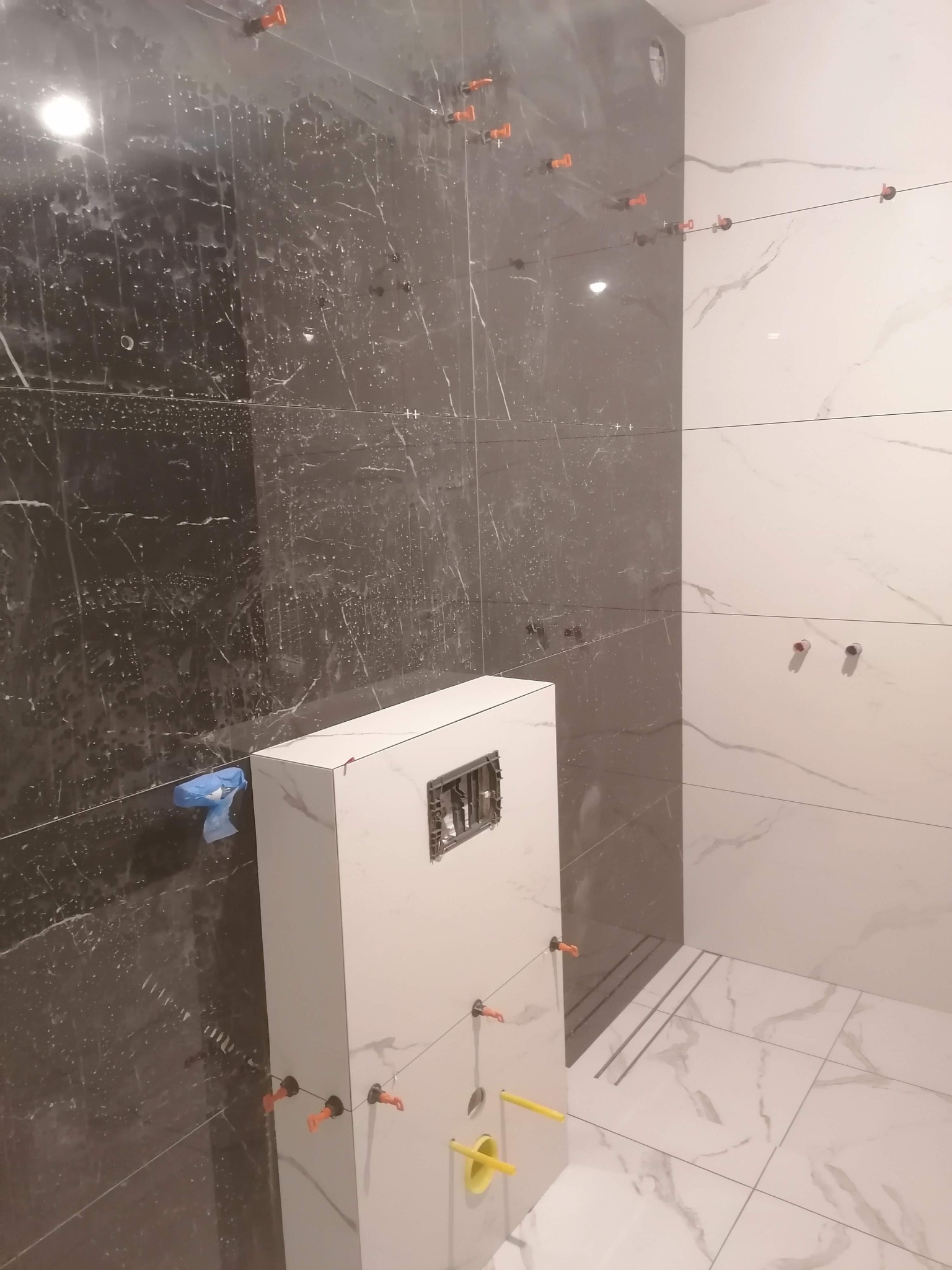 Biało-czarna łazienka w trakcie układania płytek imitujących marmur, widoczne poziomice sznurkowe, system poziomowania płytek i podtynkowy stelaż WC.