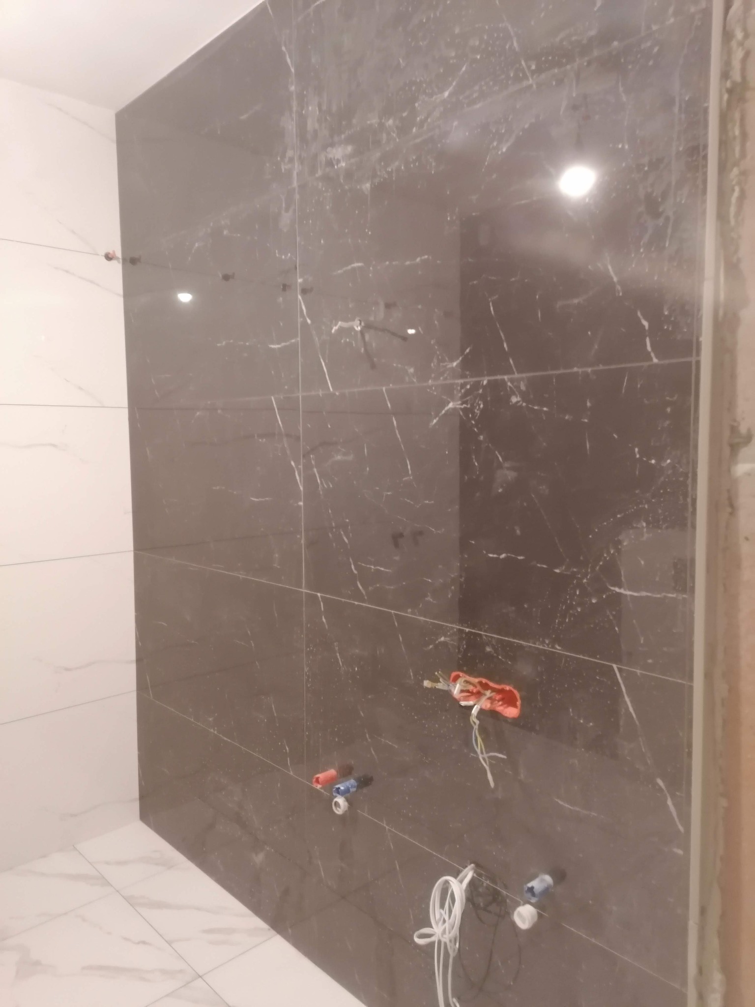 Montaż płytek imitujących marmur na ścianie, widoczne przewody elektryczne i rury wodne przed podłączeniem armatury, białe płytki podłogowe.