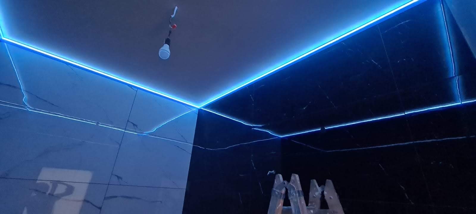 Nowoczesna łazienka z czarnymi i białymi marmurowymi płytkami, oświetlona niebieskim paskiem LED zamontowanym pod sufitem, z widoczną żarówką na kablu i fragmentem drabiny.
