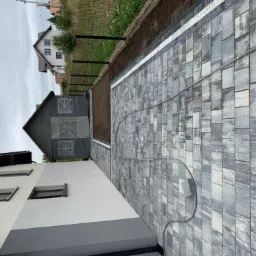 Świeżo ułożona, szara kostka brukowa o nieregularnym kształcie prowadząca od budynku do ogrodzenia, z widocznym kablem elektrycznym na powierzchni.