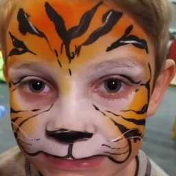 Portret chłopca z pomalowaną twarzą w tygrysa, z wyraźnymi czarnymi paskami i pomarańczowym tłem na czole i policzkach, białe akcenty wokół oczu i ust, skierowany prosto w obiektyw.