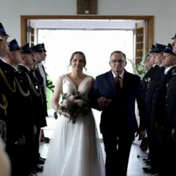 Panna młoda w białej sukni prowadzona do ołtarza przez ojca, otoczona szpalerem strażaków w galowych mundurach.