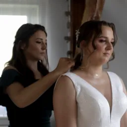 Kobieta w zielonej sukience zapina złoty naszyjnik na szyi panny młodej w białej, błyszczącej sukni ślubnej, delikatny makijaż i upięte włosy.