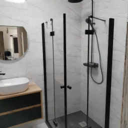 Nowoczesna łazienka z kabiną prysznicową typu walk-in z czarnymi profilami, białą umywalką nablatową na drewnianej szafce i marmurowymi płytkami na ścianach.