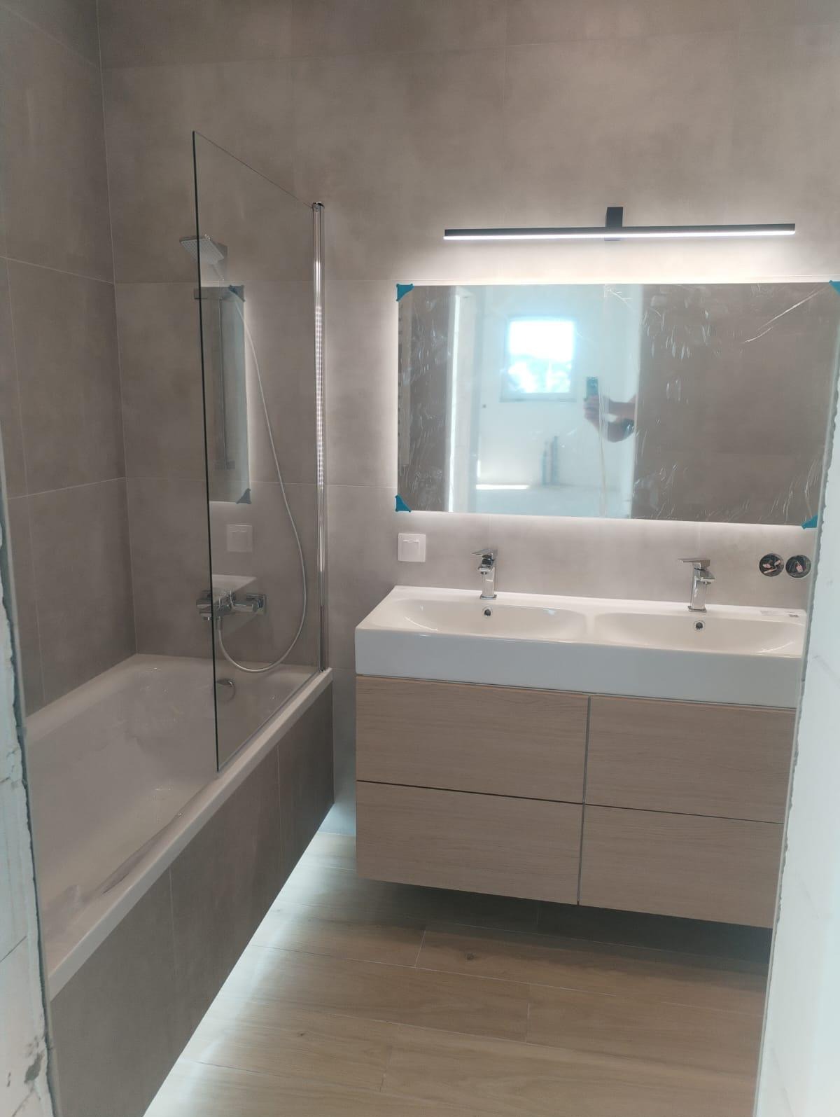 Nowoczesna łazienka z wanną za szklanym parawanem, podwójną umywalką z szafką w kolorze jasnego drewna, oświetleniem LED nad lustrem, płytki w odcieniach szarości, lustro jeszcze w folii ochronnej.