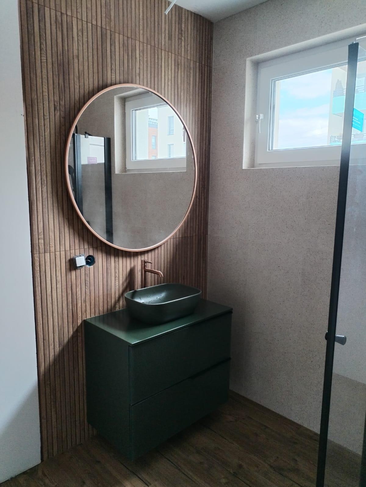 Nowoczesna łazienka z umywalką nablatową w kolorze butelkowej zieleni, okrągłym lustrem w miedzianej ramie i drewnopodobną okładziną ścienną z pionowymi żłobieniami.