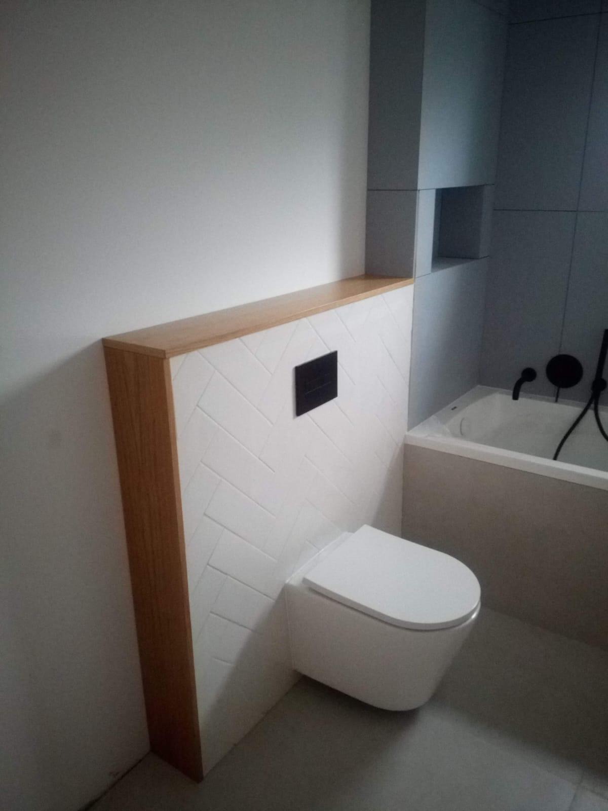 Nowoczesna toaleta podwieszana z białą armaturą i białą płytką ułożoną w jodełkę, obudowana drewnianą konstrukcją z półką, w tle wanna z czarną baterią.
