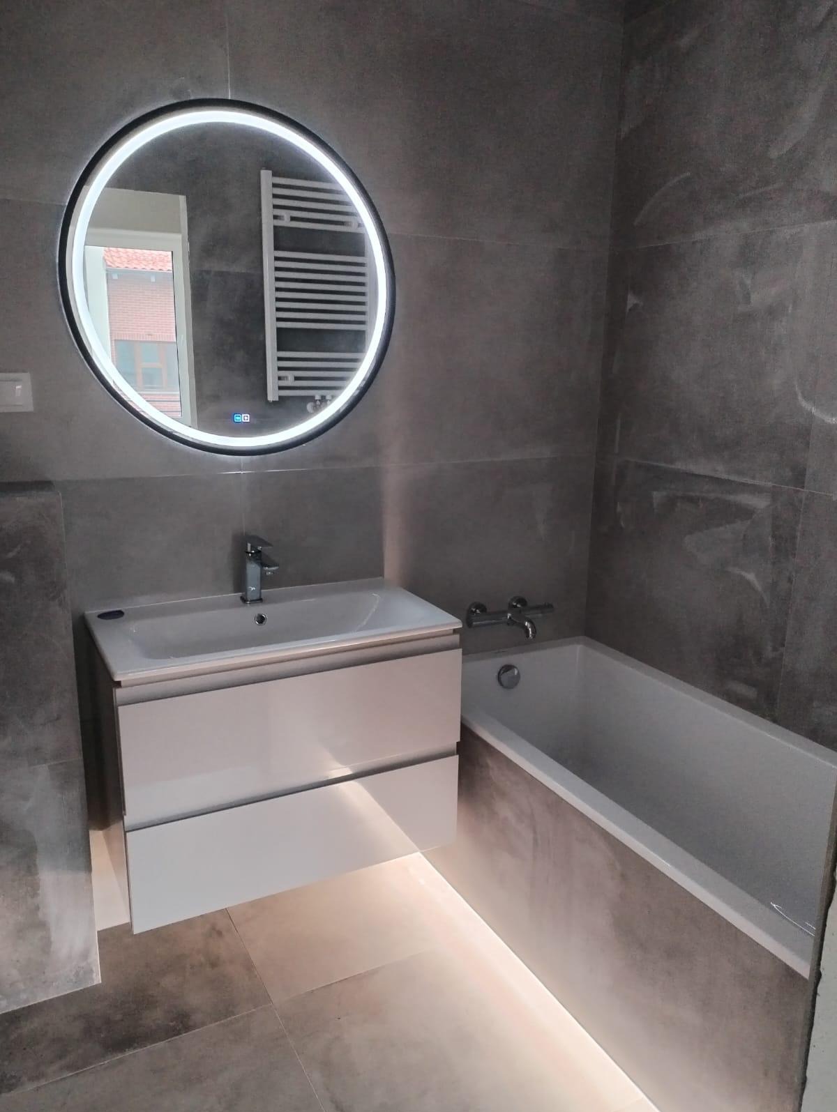 Nowoczesna łazienka w szarościach z podświetlanym lustrem i wanną, widoczne płytki imitujące kamień i biała szafka pod umywalką z subtelnym podświetleniem LED.