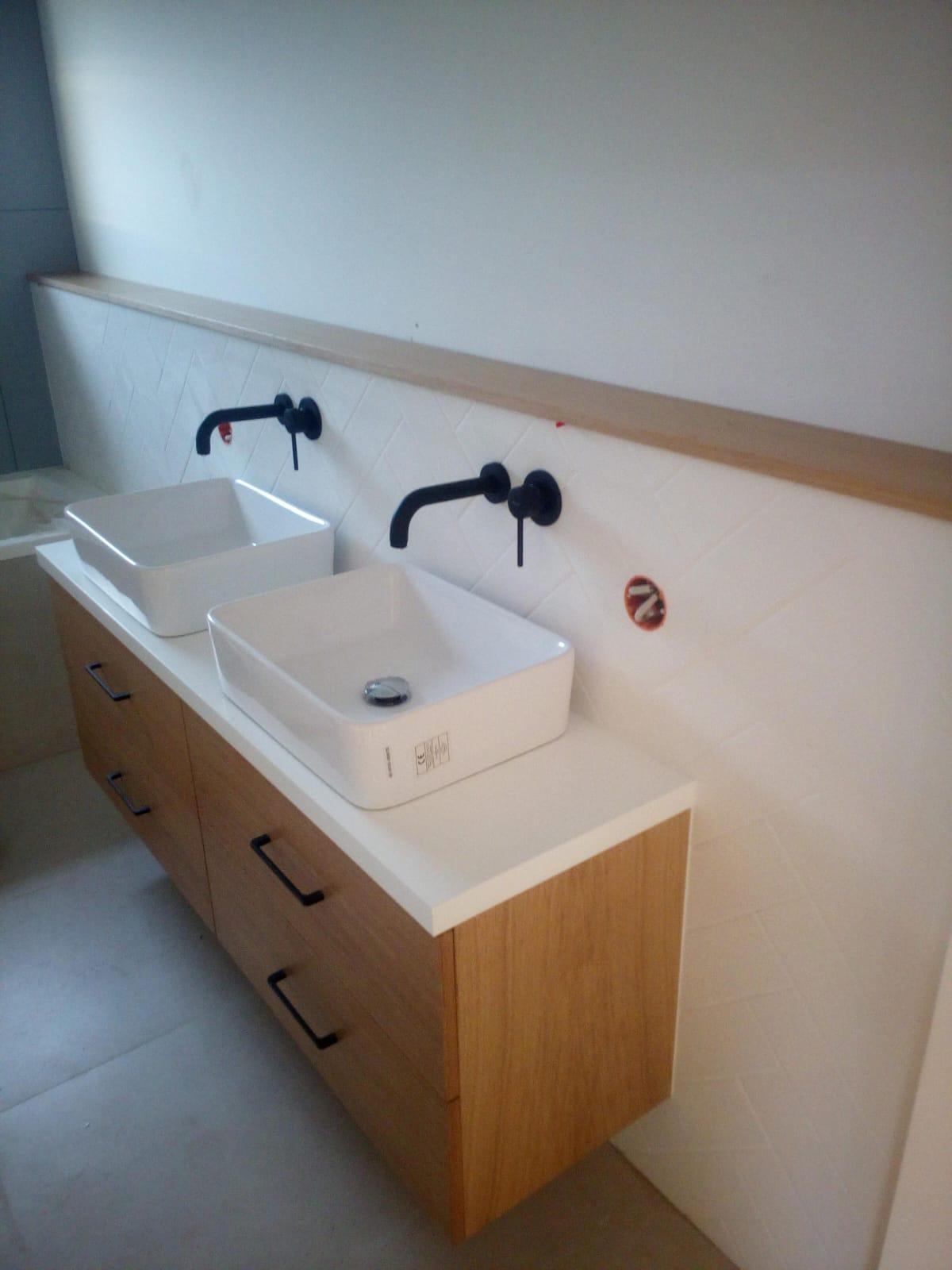 Nowoczesna łazienka z dwiema białymi umywalkami nablatowymi na szafce z drewnianymi frontami, czarne baterie umywalkowe, białe płytki ułożone w jodełkę na ścianie, widoczny fragment instalacji...