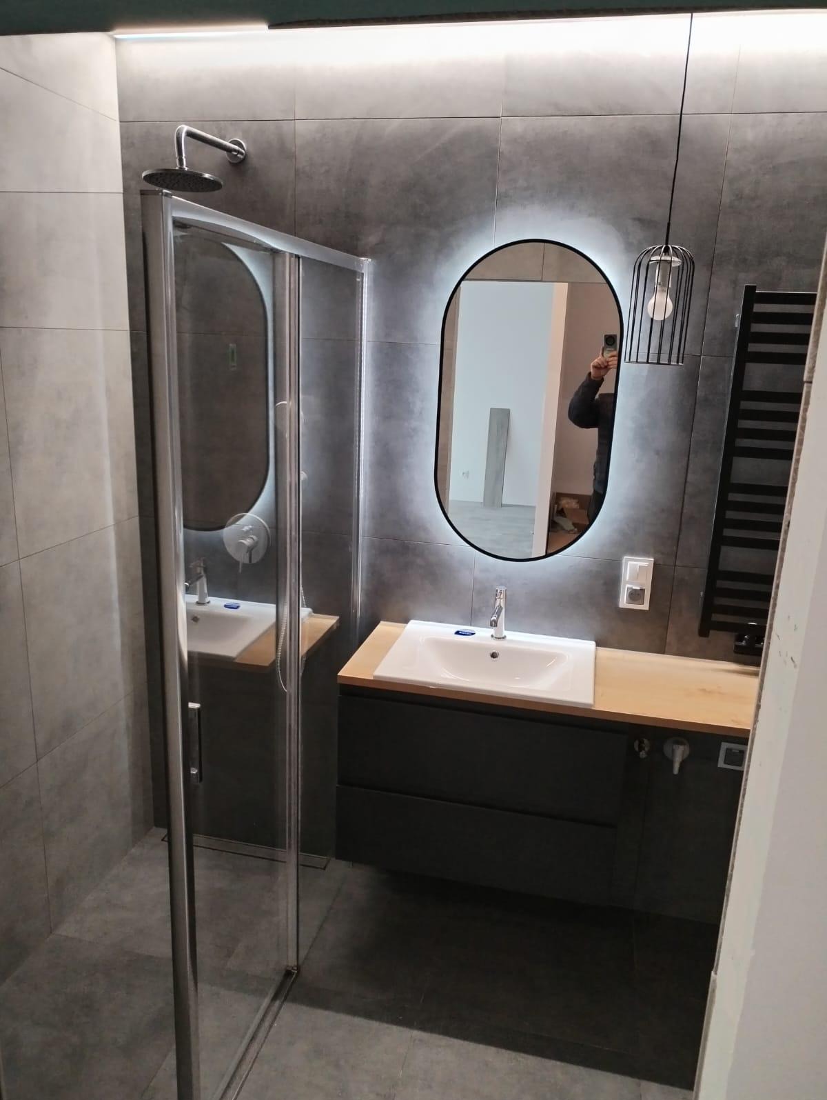 Nowoczesna łazienka z szarymi płytkami, kabiną prysznicową, umywalką nablatową z szafką i podświetlanym lustrem owalnym. W lustrze widoczne odbicie osoby robiącej zdjęcie telefonem.