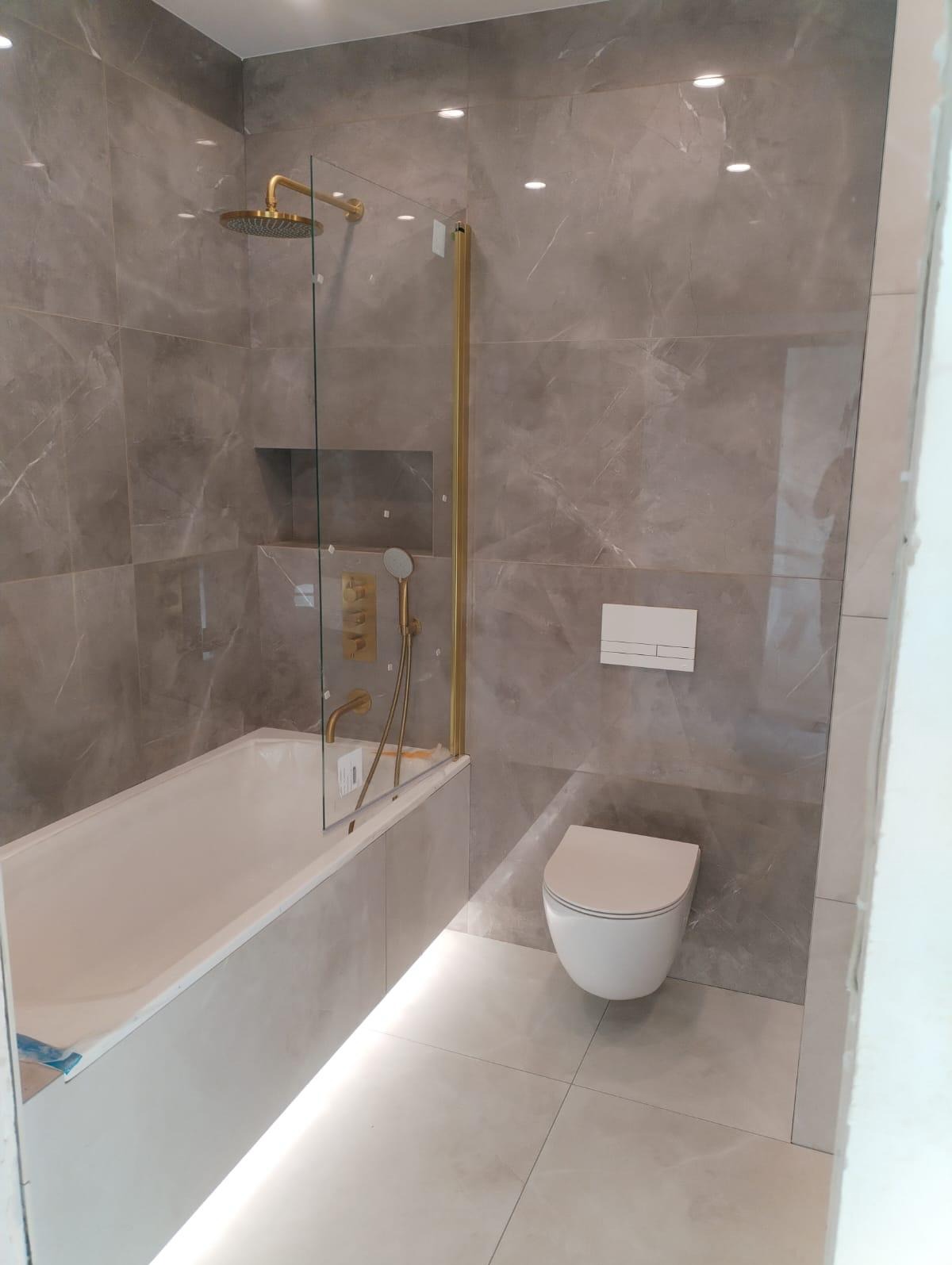 Nowoczesna łazienka z marmurowymi płytkami, kabiną prysznicową z złotymi elementami, podświetlaną wanną i podwieszaną toaletą.