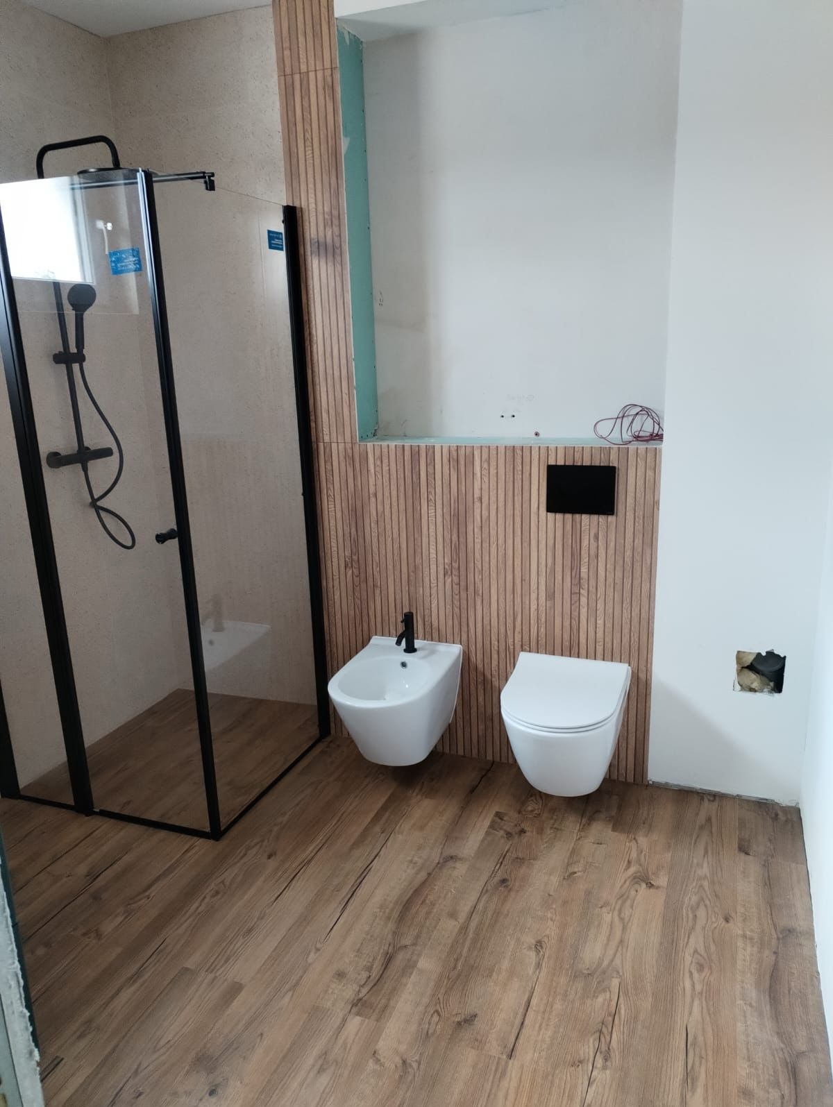 Wnętrze łazienki w trakcie prac: kabina prysznicowa z czarnymi ramami, wiszący bidet z czarną armaturą i toaleta, drewnopodobne panele na ścianie, widoczne niedokończone fragmenty ściany.