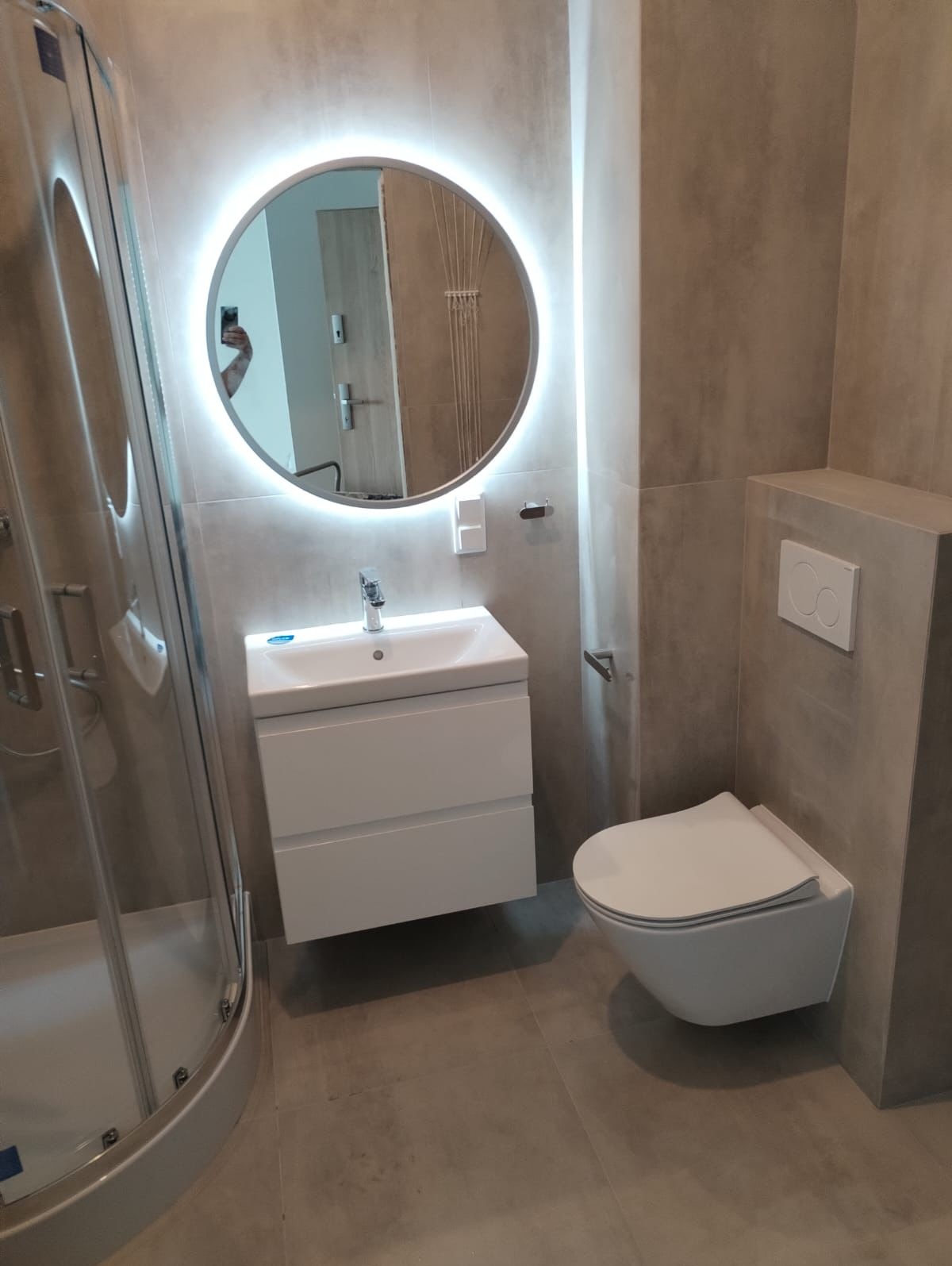 Nowoczesna łazienka z kabiną prysznicową, podwieszaną toaletą i okrągłym lustrem z podświetleniem LED, szare płytki imitujące beton.