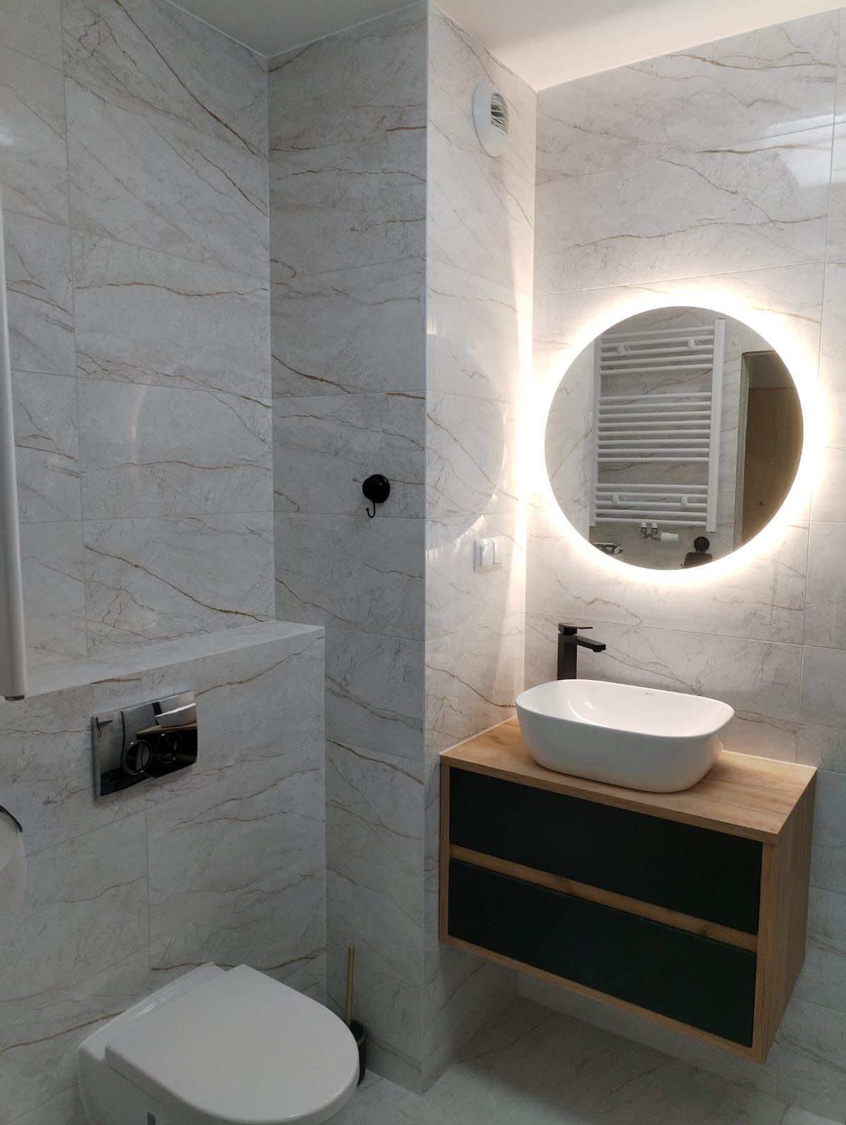 Nowoczesna łazienka z marmurowymi płytkami, okrągłym lustrem z podświetleniem LED, białą umywalką nablatową na szafce z drewnianym blatem i czarnymi frontami oraz wiszącą toaletą.